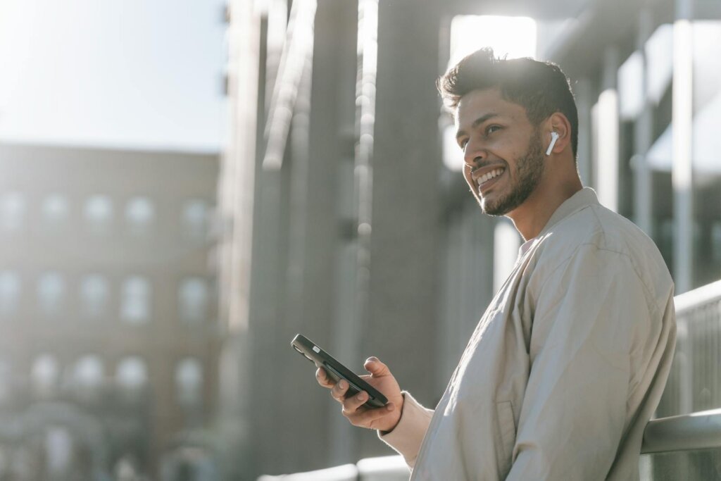 Autoridade digital - Side view of cheerful young bearded Hispanic male in TWS earbuds using mobile phone while standing near modern urban building in sunny day
